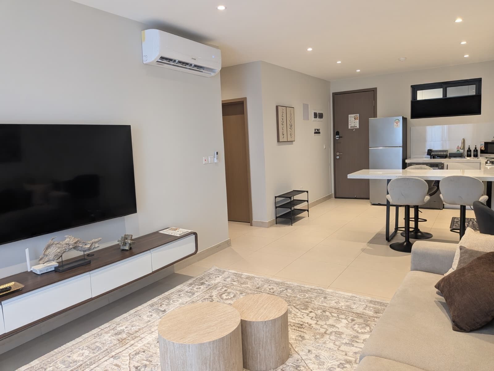 A modern living room with a flat-screen TV, a sofa, a coffee table, and a kitchen area with a refrigerator and a dining table.
