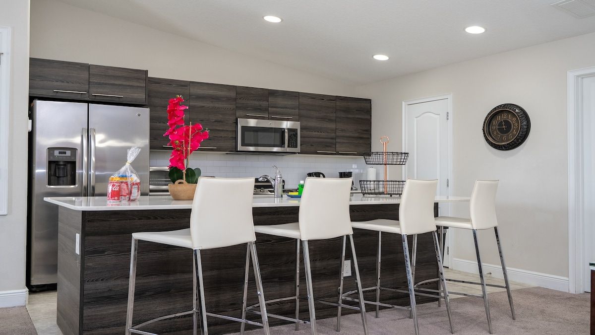 A modern kitchen with a central island, bar stools, a refrigerator, a microwave, and a clock on the wall.