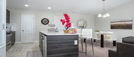 Una cocina moderna con una isla central, un comedor con una mesa y sillas blancas, y una sala de estar con sofá.