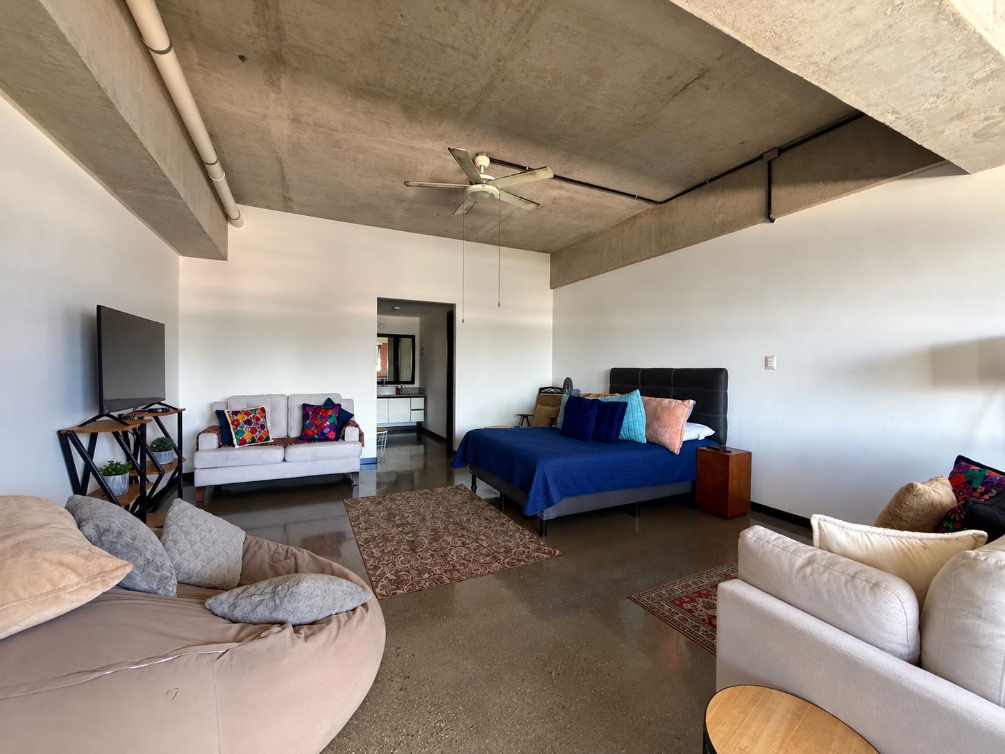 A modern living room with a sofa, a bed with a blue bedspread, a ceiling fan, and a television.