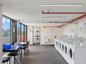 A laundry facility with multiple washing machines, a table with chairs, and a view through large windows. - [New] Ocean View 2 bedroom 2 bath in Waikiki (Honolulu)