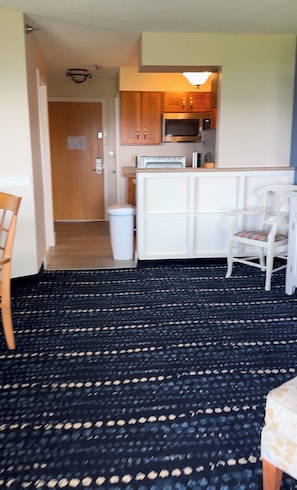 A room with a patterned carpet, a white chair, a small table, and a kitchenette area with wooden cabinets and a microwave.