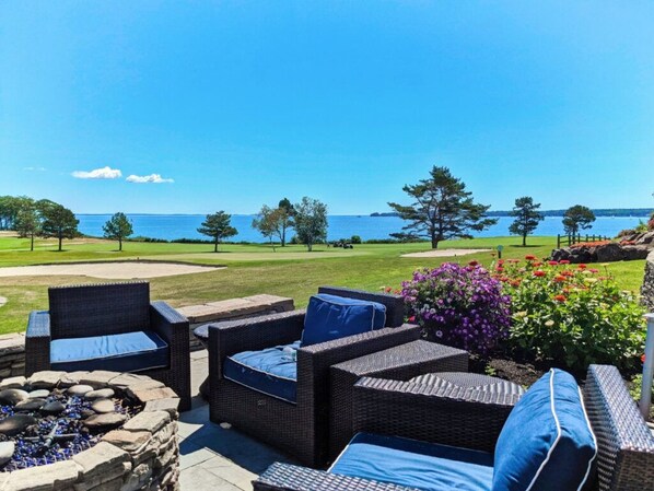 An outdoor seating area with wicker furniture and a fire pit, overlooking a golf course and the ocean.