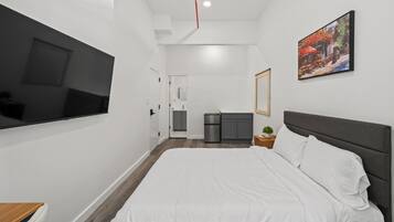 Une chambre d’hôtel moderne dotée d’un grand lit, d’un téléviseur à écran plat fixé au mur, d’une petite kitchenette et d’un tableau décoratif.