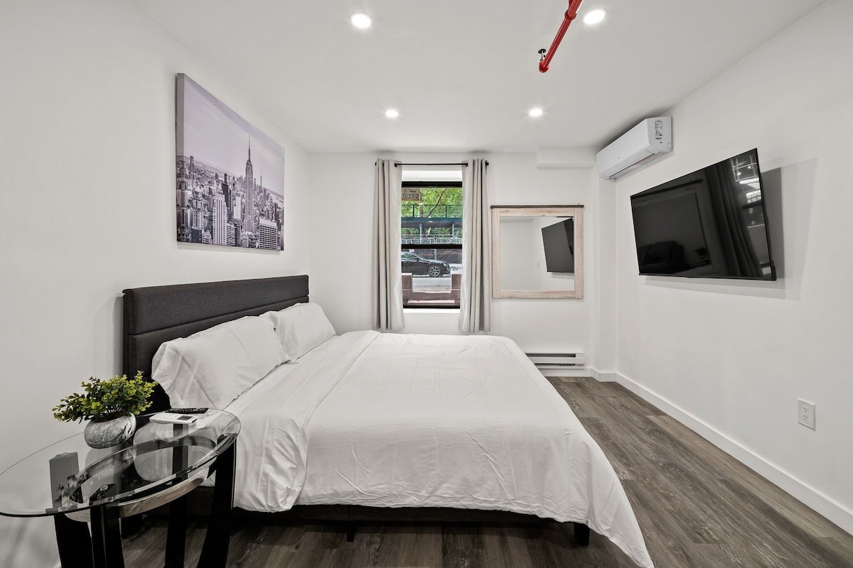 A modern bedroom with a bed, a glass side table, a cityscape painting, an air conditioning unit, and a TV mounted on the wall.