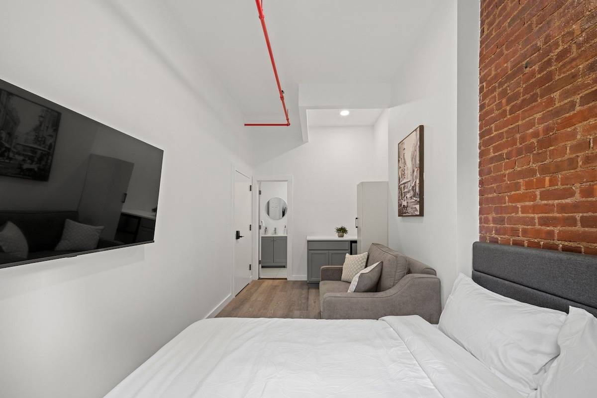 A modern bedroom with a bed, a grey sofa, a mirror, and a brick wall.