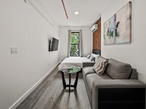 A modern hotel room with a bed, a sofa, a glass coffee table, a TV mounted on the wall, and a window with a view of trees.