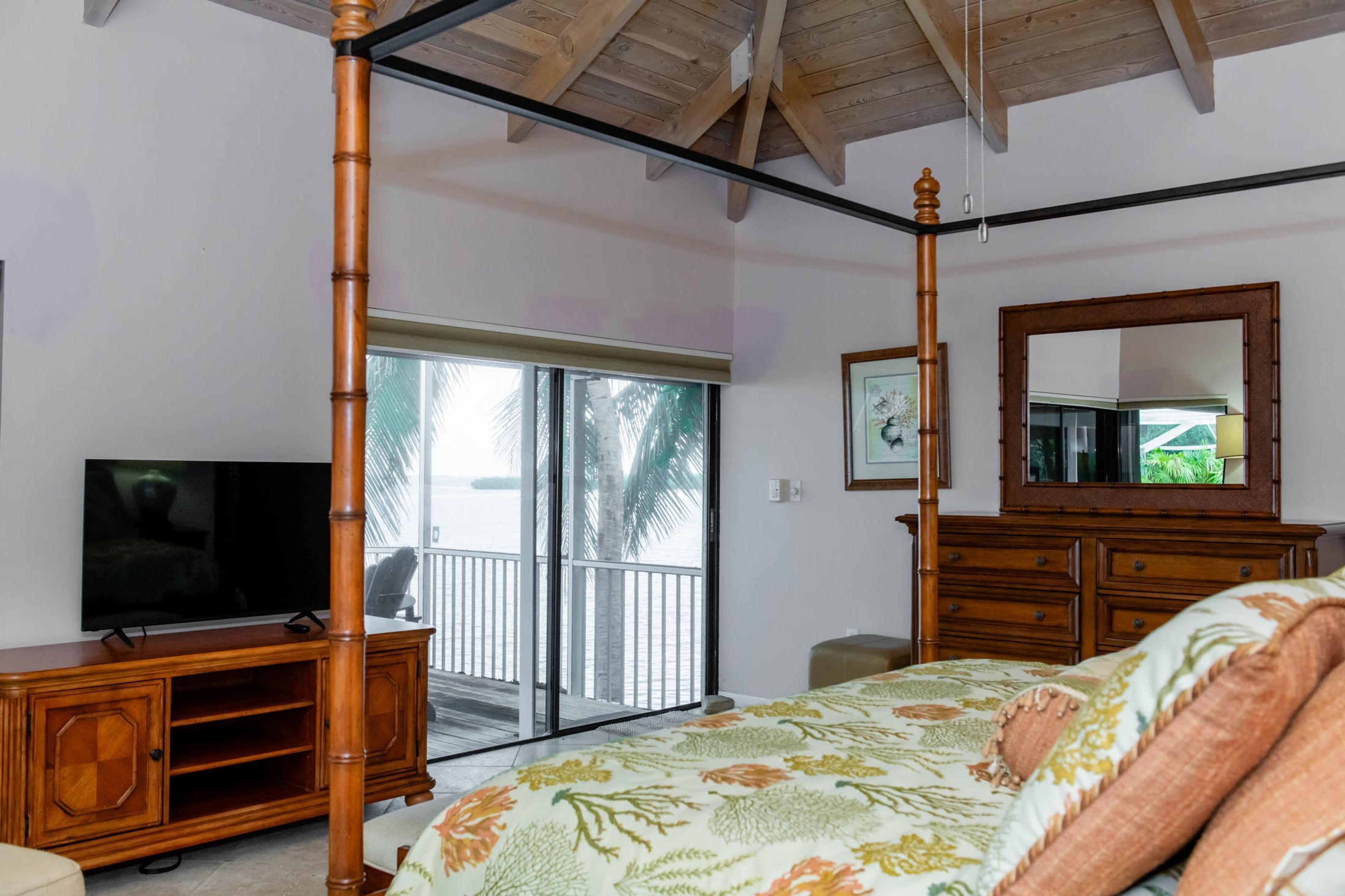 A bedroom with a four-poster bed, a television on a wooden cabinet, a large mirror, and a sliding glass door leading to a balcony.