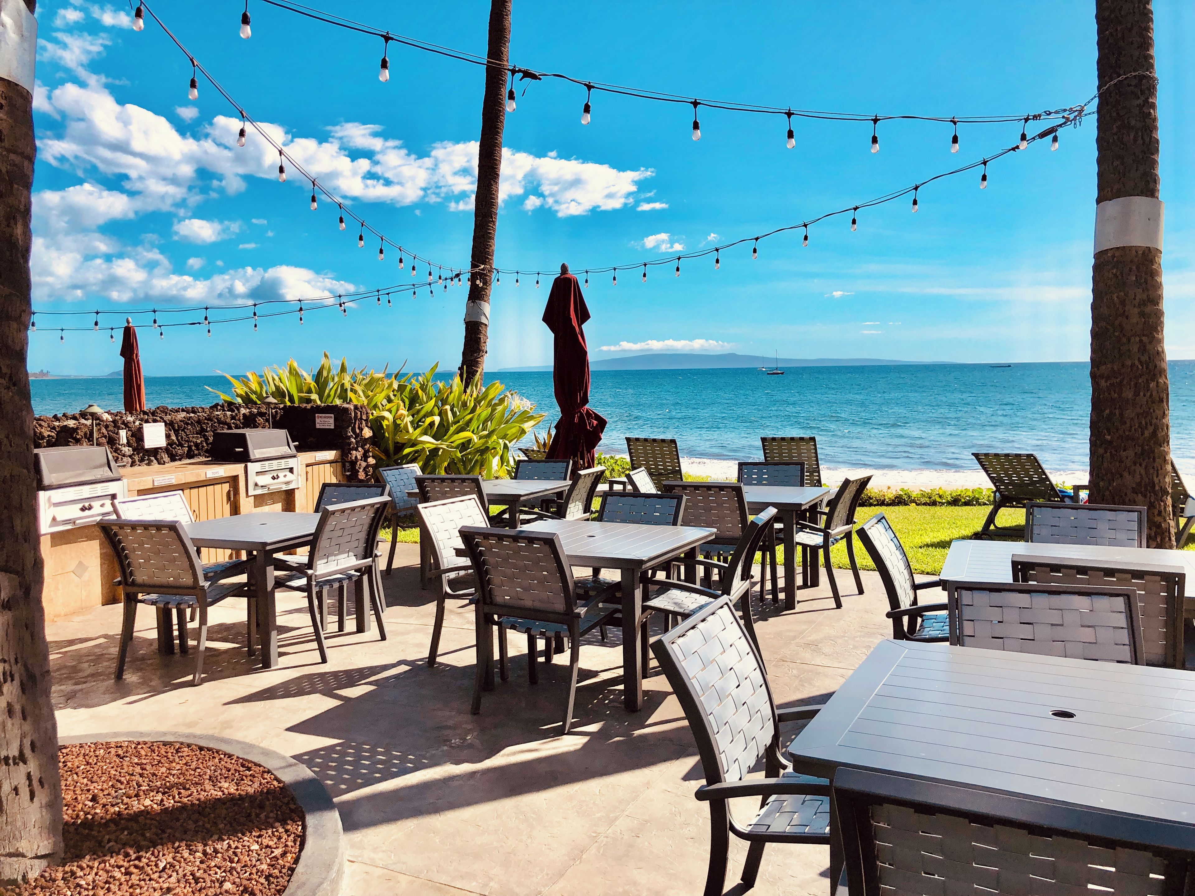 An outdoor seating area with tables and chairs, a grill, and string lights against a backdrop of the ocean and sky.