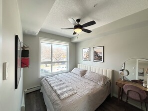 A bedroom with a bed, a desk, a chair, a ceiling fan, and a window with a view of the outside.