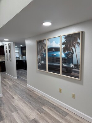 A hallway with a wooden floor, a kitchen area in the background, and a wall-mounted picture of a vintage car.