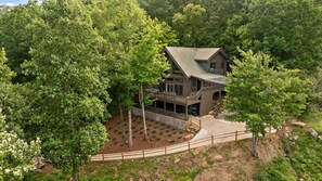 A two-story log cabin with a balcony and a covered porch, surrounded by dense forest.