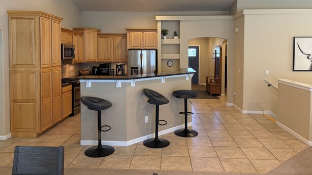 A kitchen with wooden cabinets, a stainless steel refrigerator, a microwave, and a coffee maker. There are three black bar stools and a small island with a sink.