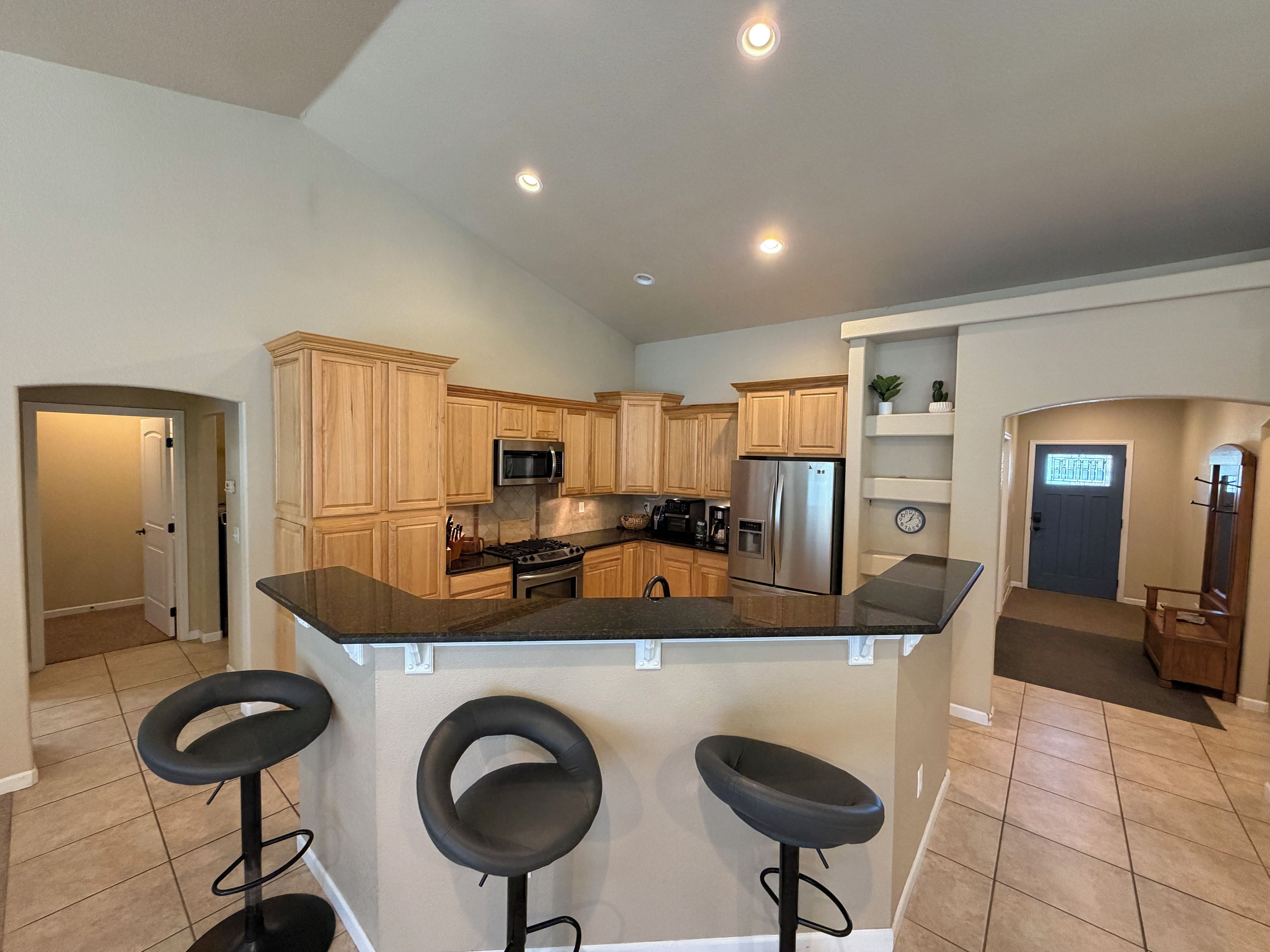 A kitchen with wooden cabinets, a stainless steel refrigerator, a microwave, and a gas stove. There are three black bar stools in front of the counter.