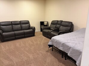 A room with a beige carpet, two leather sofas, a black TV stand, and a bed with a white quilt.