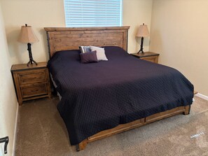 A bedroom with a wooden headboard, a dark quilted bedspread, two bedside tables with lamps, and a window with blinds.