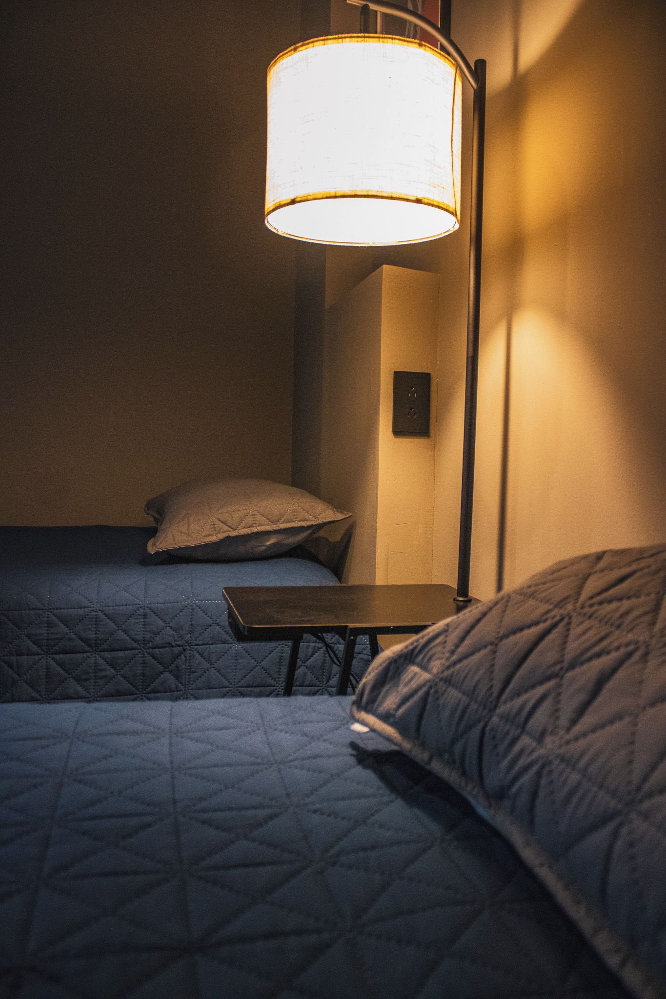 Une pièce faiblement éclairée, avec deux lits, une petite table et une lampe fixée au mur.