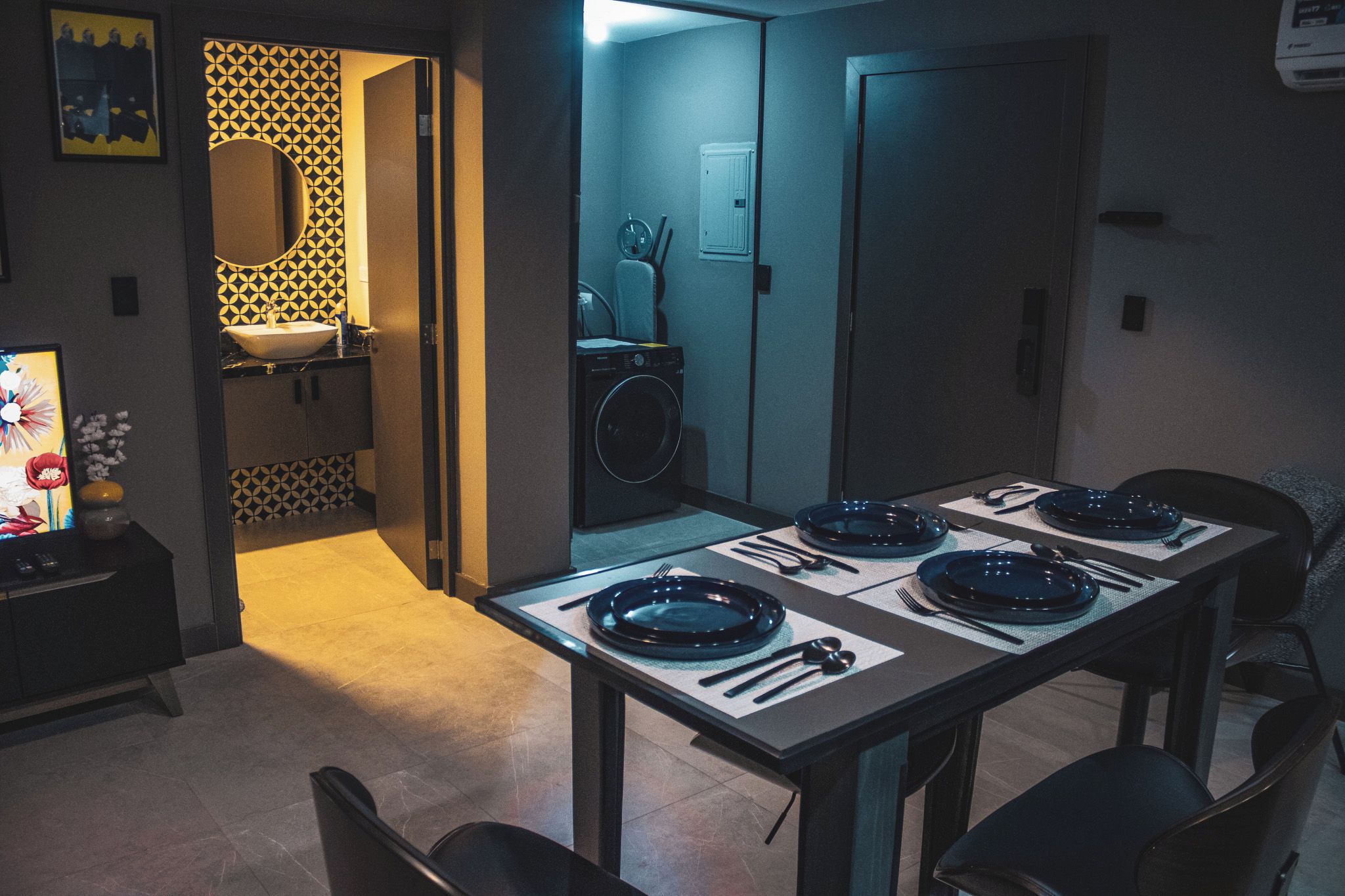 Un comedor con una mesa dispuesta para cuatro personas, una lavadora y un baño visible a través de una puerta abierta.