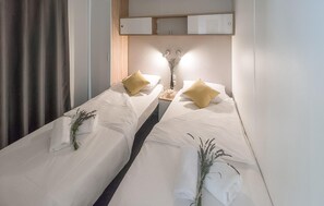 Una camera d'albergo con due letti, ognuno con una coperta bianca e un cuscino giallo. Tra i letti c'è un tavolino con un vaso di fiori secchi.