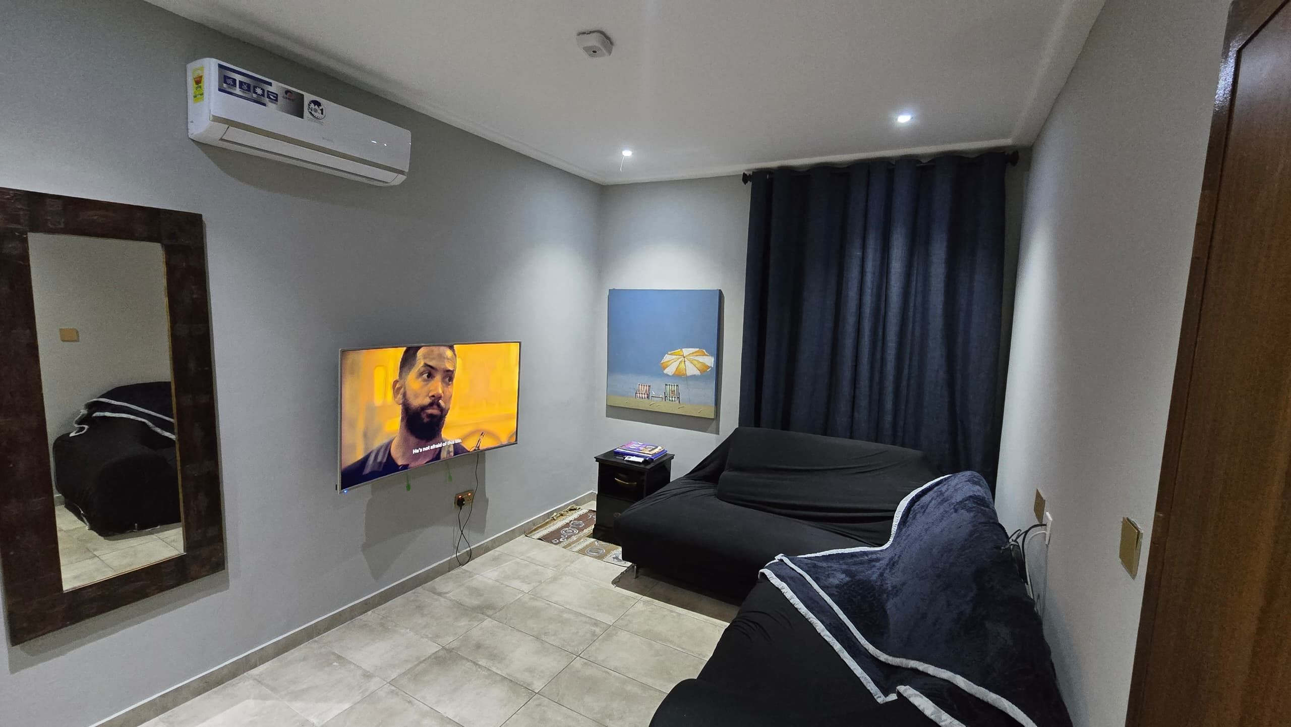 Un espace de vie compact comprenant un téléviseur à écran plat fixé au mur, un canapé, une petite table avec une lampe et une fenêtre avec des rideaux.