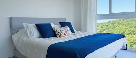 Cama com roupa de cama branca e azul, cabeceira cinza e vista para uma paisagem verde pela janela.