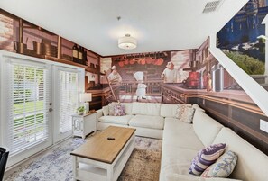 A living room with a large mural, a white sofa, a wooden coffee table, and a patterned rug. - 4BR Themed Home W/Resort Pool Near Disney (Kissimmee)
