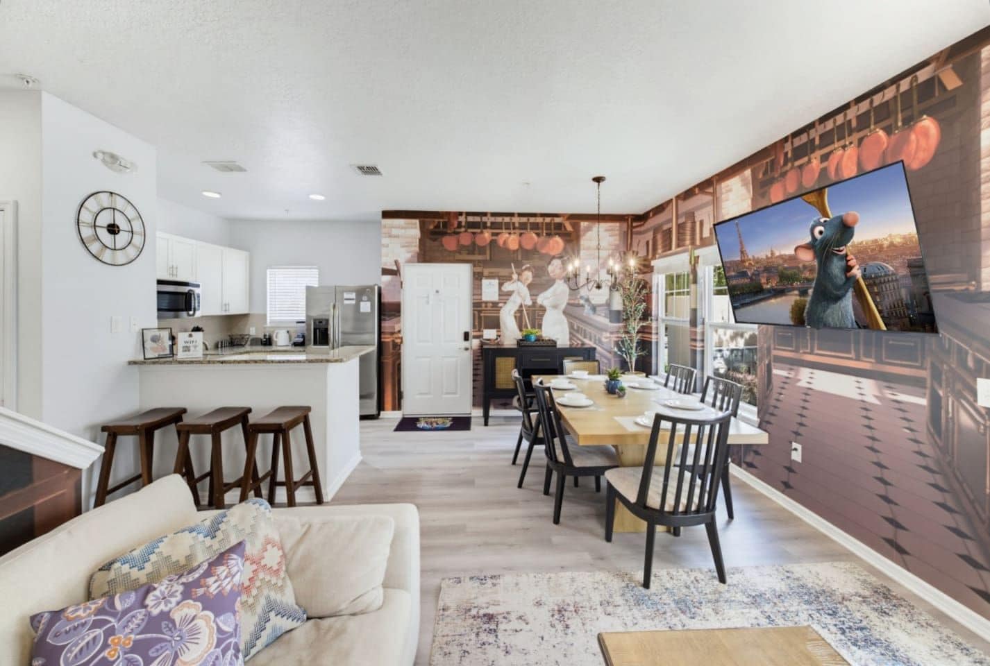 Un salon moderne avec un canapé, un coin repas comprenant une table et des chaises, une cuisine équipée et une grande fresque murale.