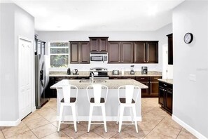 A modern kitchen with dark wood cabinets, a stainless steel refrigerator, a microwave, and a granite countertop. There are three white bar stools at the island. - Lakefront 5BR Family Resort Pool Home Near Disney (Kissimmee)