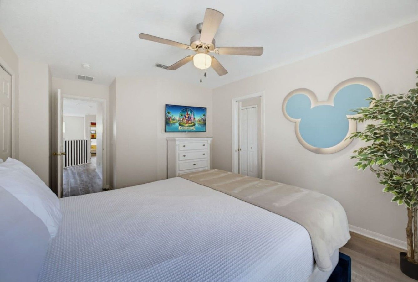 A bedroom with a bed, a ceiling fan, a dresser, a Mickey Mouse wall decoration, and a potted plant.