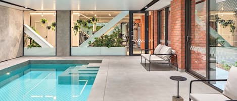 Ein moderner Innenpoolbereich mit einem klaren, blauen Pool, einer Treppe zu einer erhöhten Ebene und einer Sitzecke mit einer Couch und Sesseln.