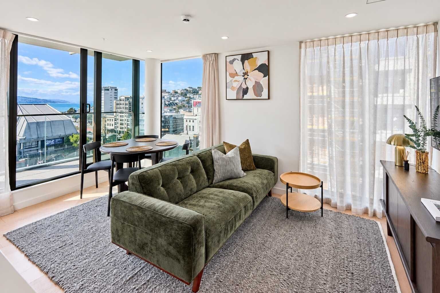 A modern living room with a green sofa, a dining table with chairs, and a city view through large windows.