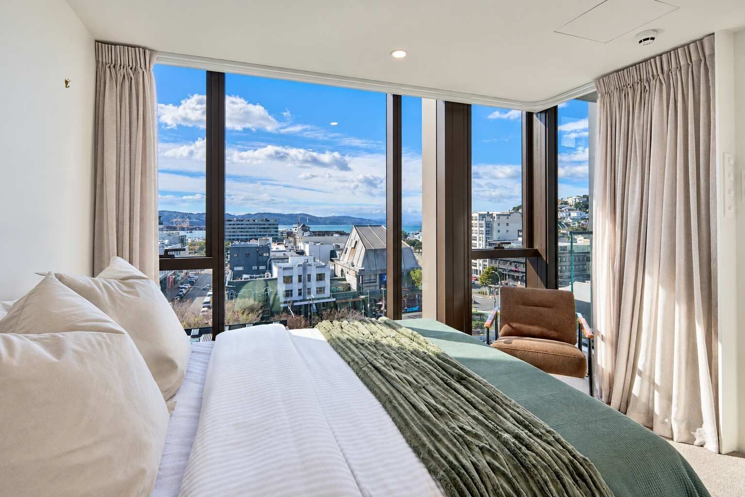A hotel room with a large bed, a chair, and a view of a cityscape through large windows.