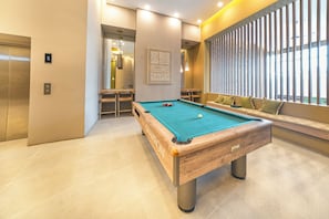Une table de billard moderne dans une pièce bien éclairée, avec un canapé et une vue sur l’extérieur grâce à de grandes fenêtres.
