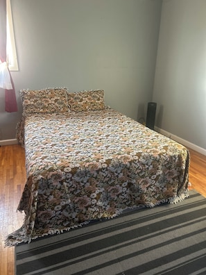 A bed with a floral bedspread, a striped rug, and a speaker in a room with wooden flooring and a plain wall.
