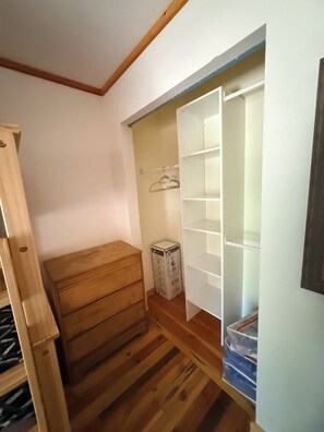 A small room with a wooden dresser, a closet with open shelves, and a trash bin.