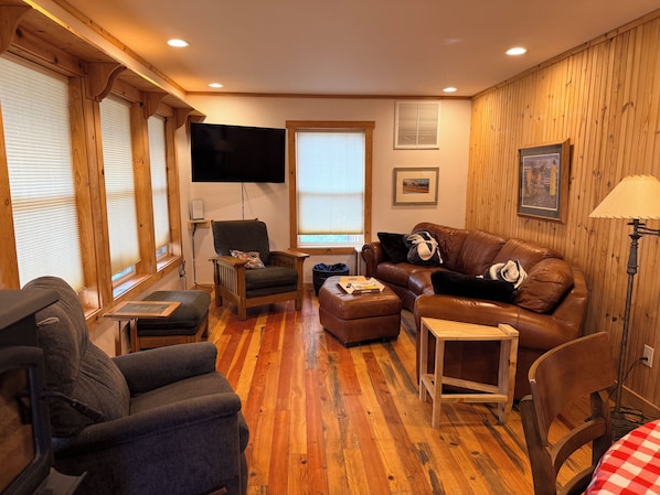A cozy living room with a brown leather sofa, a wooden armchair, a flat-screen TV mounted on the wall, and a wooden floor.