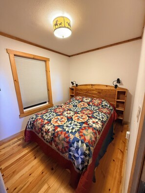 A bedroom with a wooden headboard, a patterned quilt, a window with blinds, and a ceiling light fixture.