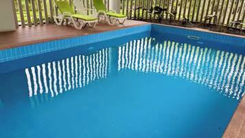 Une piscine dont le bord est recouvert de carrelage, entourée d’une terrasse en bois et de chaises longues vertes.