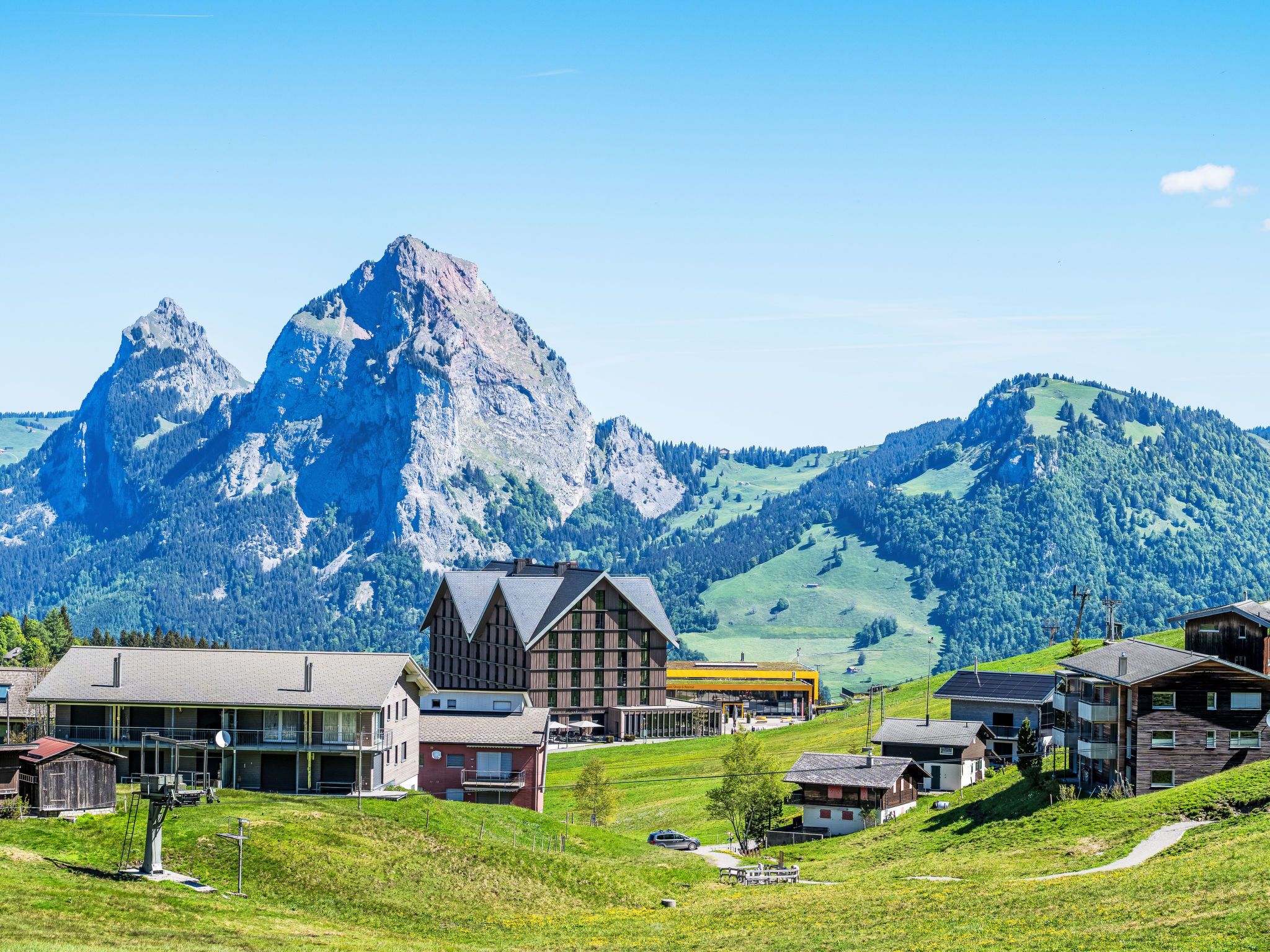 Ein Bergdorf mit traditionellen Holzhäusern, einem modernen Gebäude und einem markanten Berggipfel im Hintergrund.