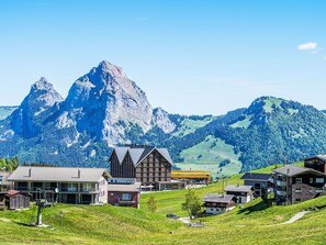 Ein Bergdorf mit traditionellen Holzhäusern, einem modernen Gebäude und einem markanten Berggipfel im Hintergrund.
