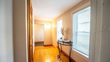 Un pasillo con piso de madera, una puerta a la izquierda, un aplique en la pared y una ventana con persianas a la derecha.