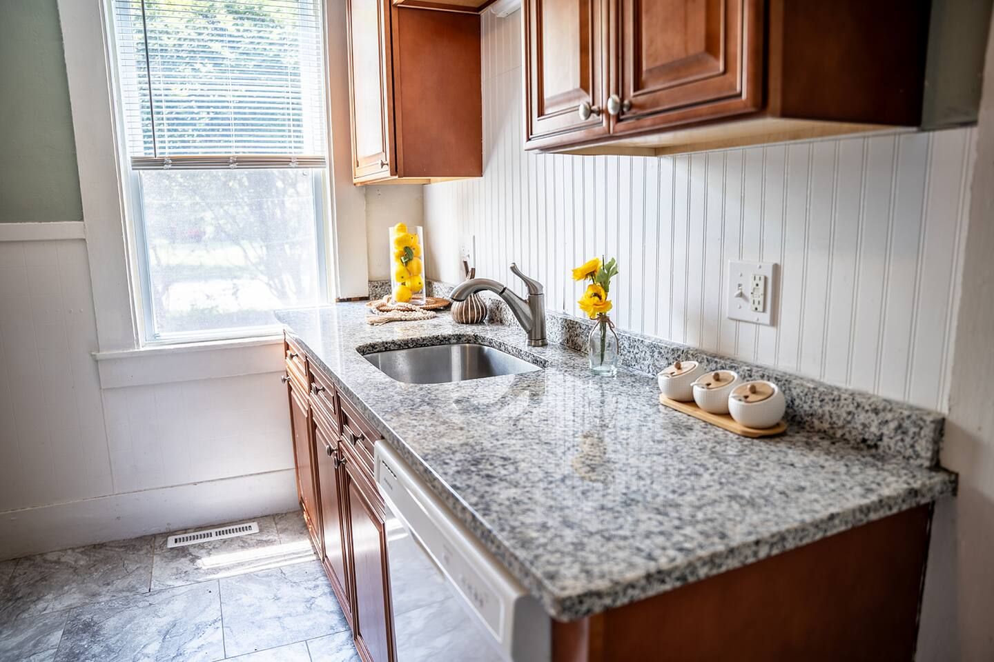 A kitchen with granite countertops, a stainless steel sink, wooden cabinets, and a window with blinds.