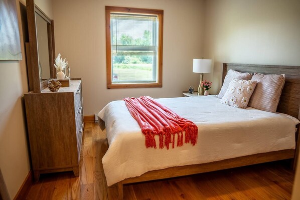 A bedroom with a large bed, a nightstand with a lamp, a dresser, and a window with blinds.