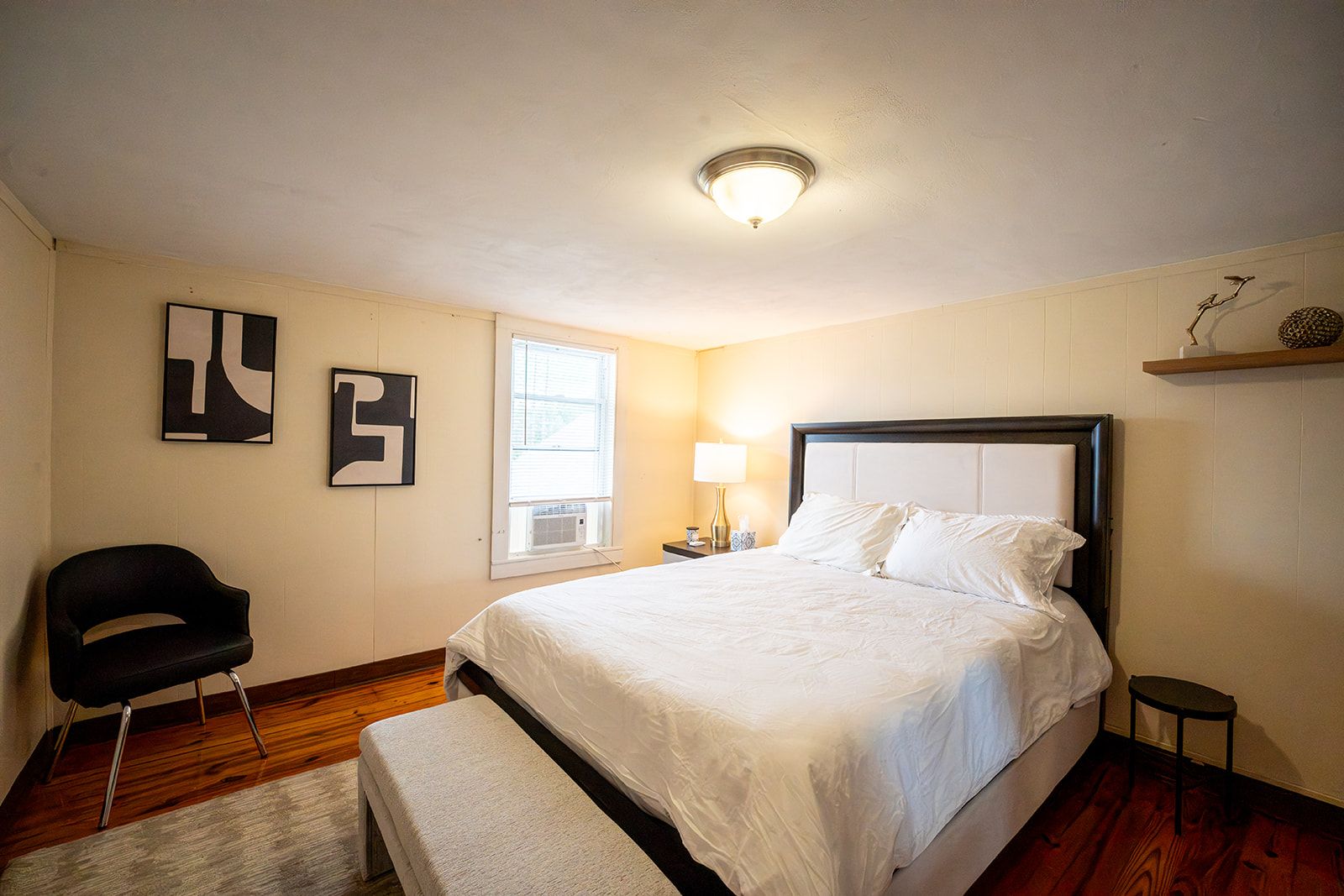 A bedroom with a bed, a chair, a bench, a nightstand, and a wall-mounted shelf.