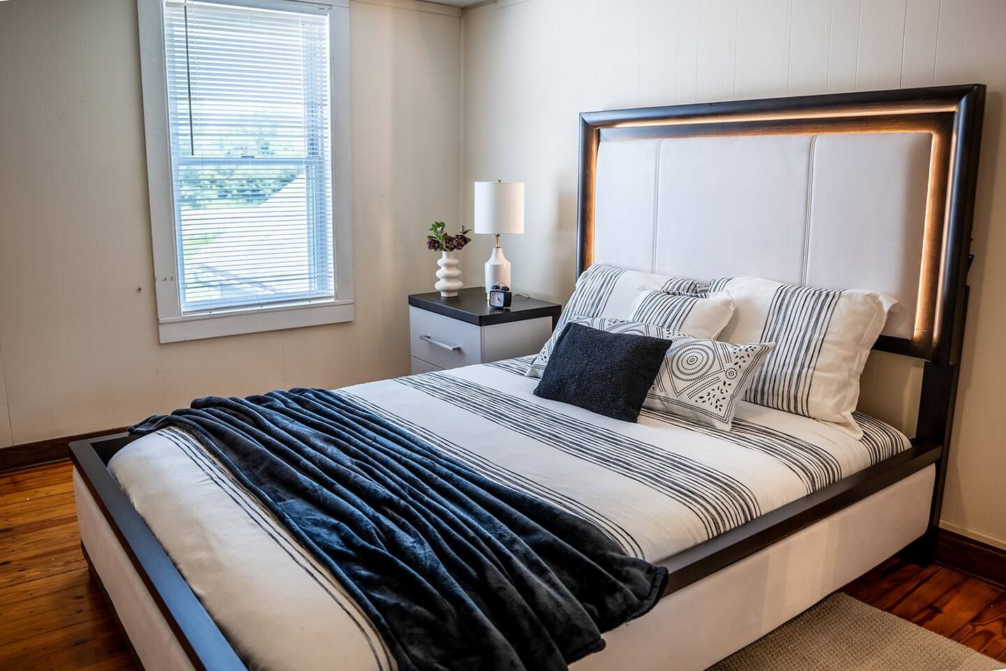 Un dormitorio moderno con cama, mesita de noche, lámpara y ventana.