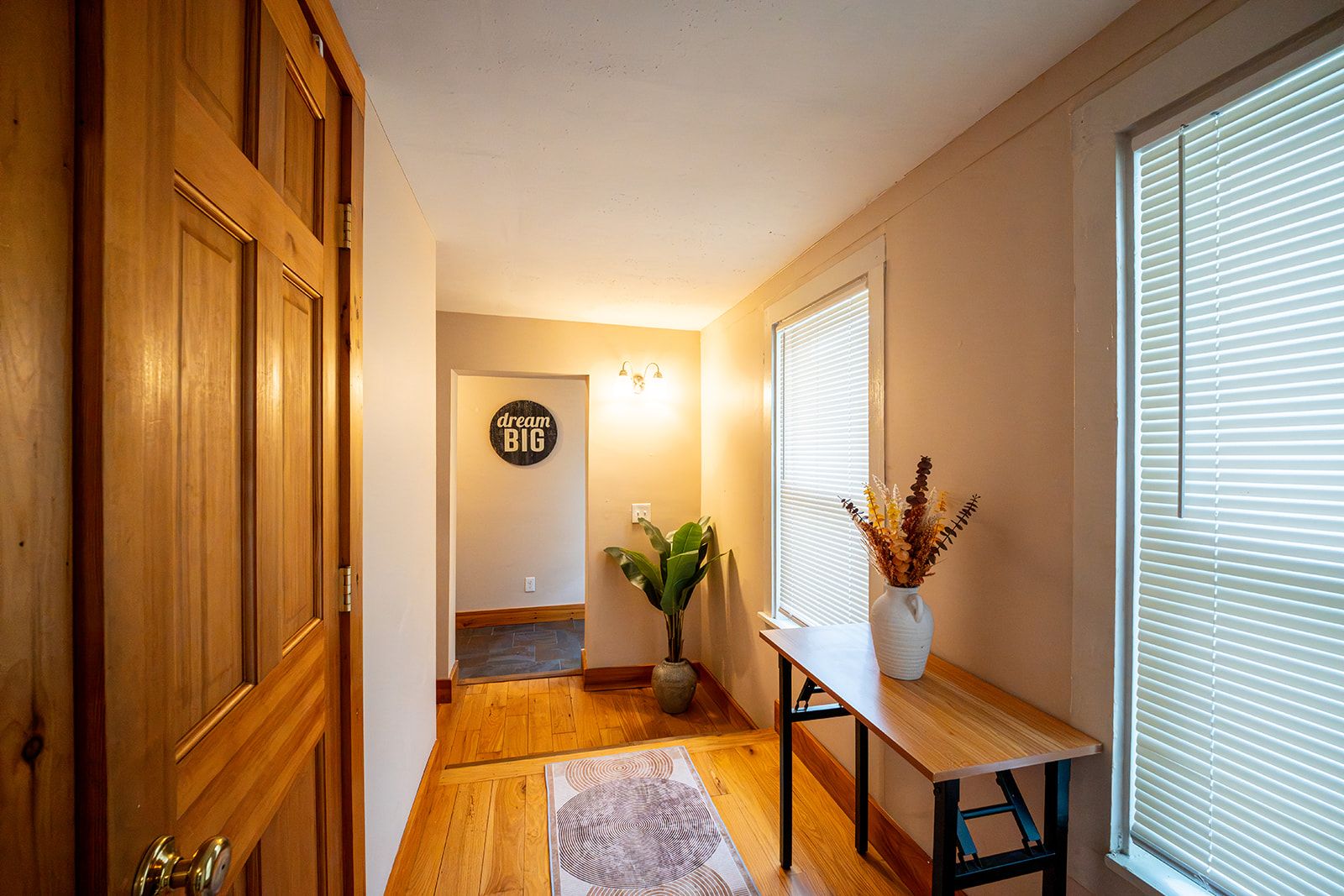 Un couloir avec des portes en bois, une enseigne fixée au mur, une plante en pot, une table avec un vase et une fenêtre avec des stores.