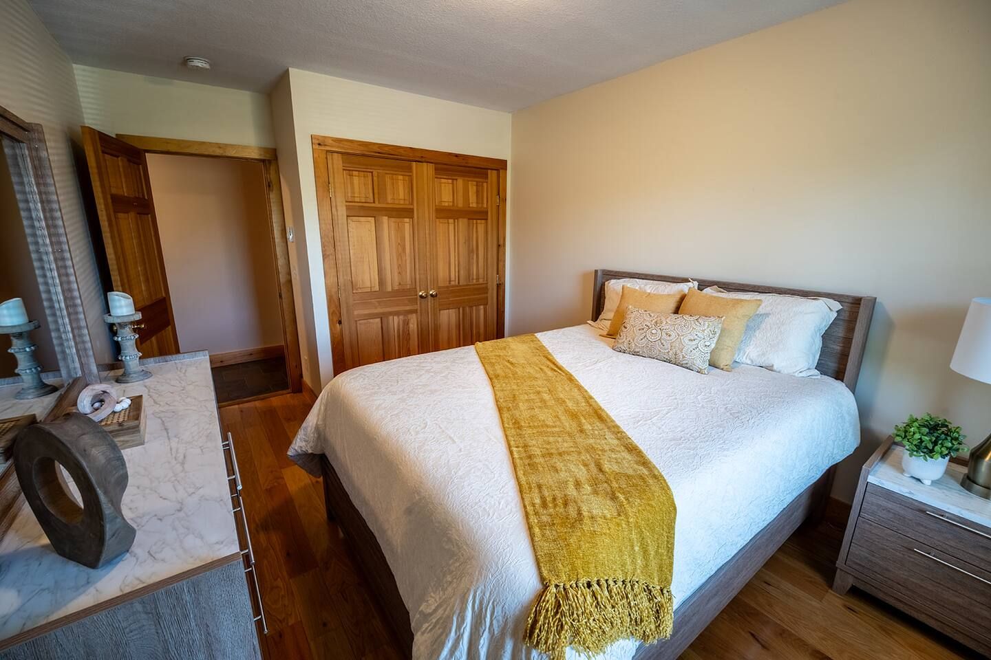 Quarto com cama, guarda-roupa de madeira, mesa de cabeceira e uma pequena planta.