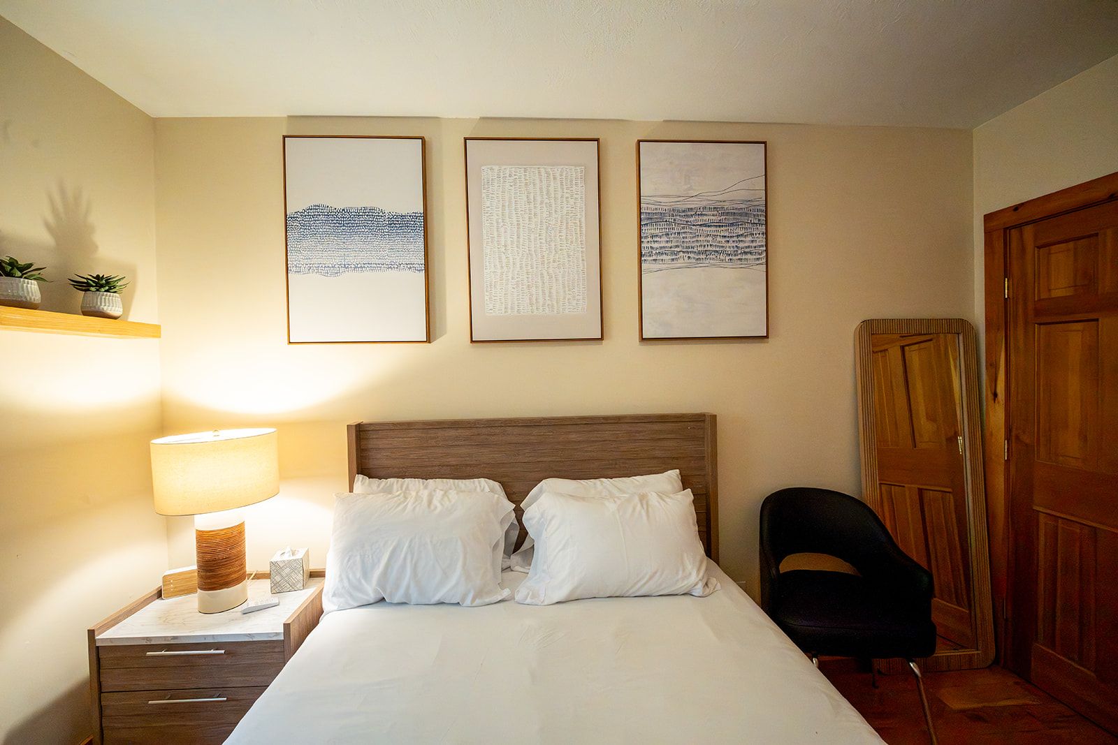 Habitación de hotel con cama, mesita de noche, lámpara, silla y tres cuadros en la pared.