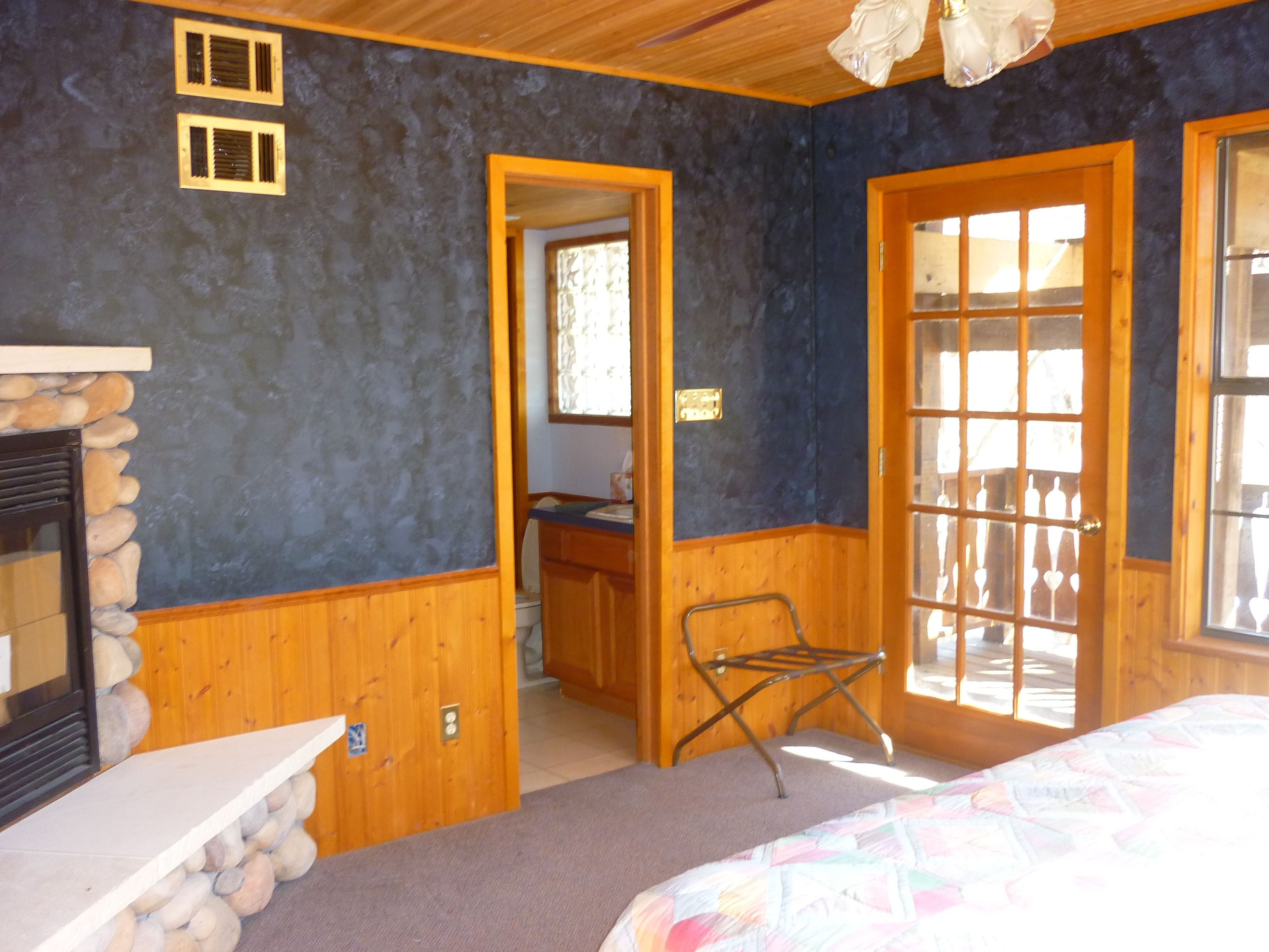 A room with a stone fireplace, wooden walls, a bed with a colorful quilt, and a door leading to an outdoor area.