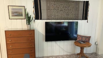 Una habitación con una televisión de pantalla plana montada en la pared, un aparador de madera, una mesita redonda con un cojín y una alfombra estampada en el suelo.